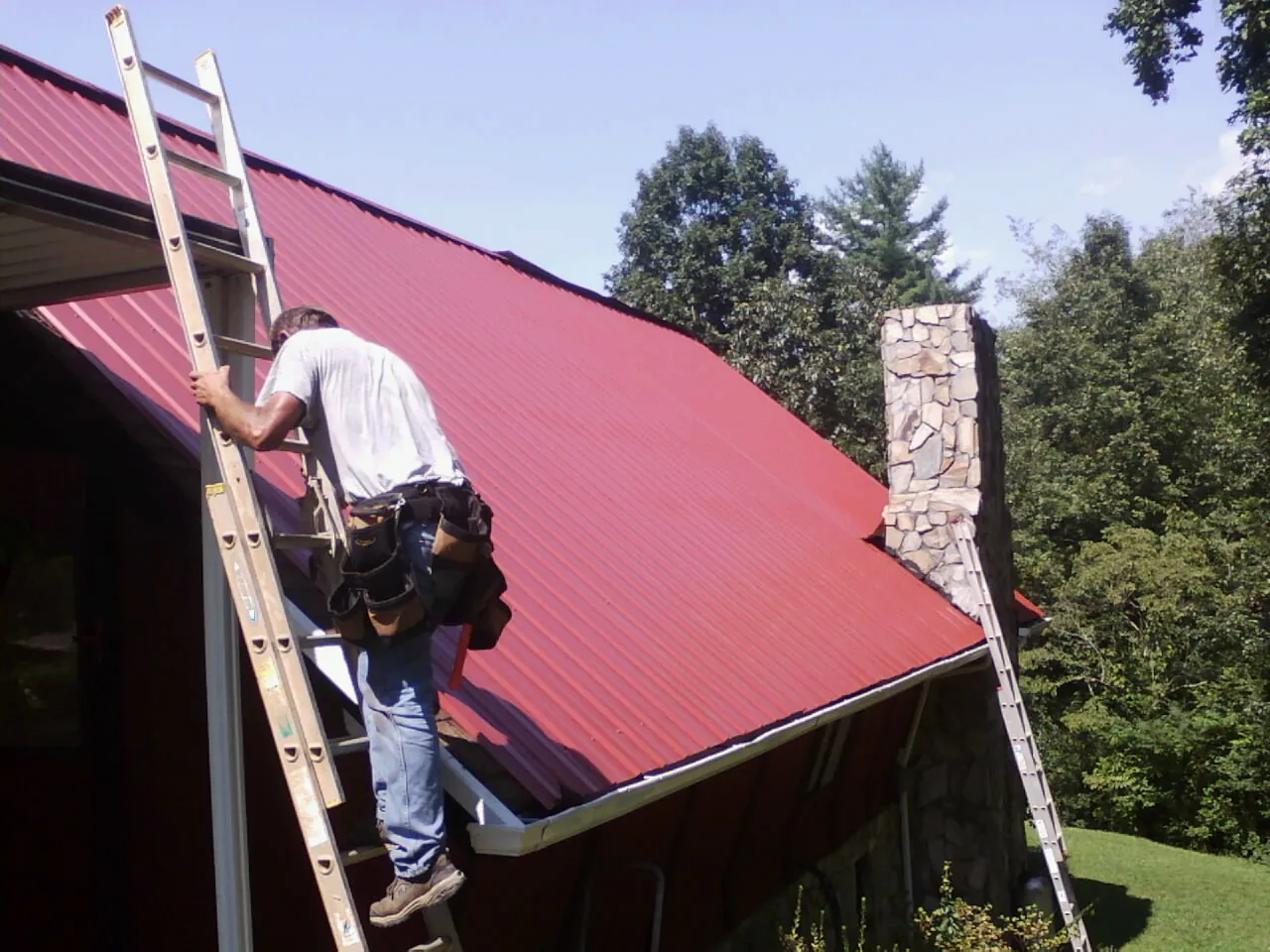 Roofer on a ladder beside a red metal roof and stone chimney — Soffit & Fascia Repair by Roofing Brothers in Newburg